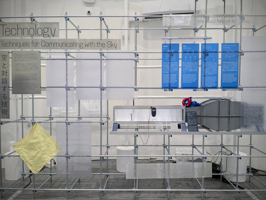 Exhibition scene of "Let's imagine the next generation of teru-teru bōzu, together!" technology display. Inside a small wind tunnel, the model of the Offshore Curtain shows its effect in reducing water vapor inflow (photo center-right). The Offshore Curtain panel is the second from the right among the blue panels. The Offshore Curtain material is displayed at the lower left of the photo.